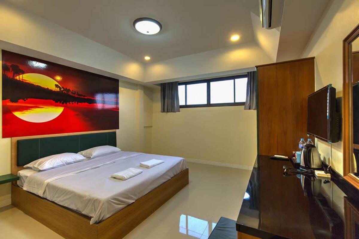 A modern hotel room with a large bed, a flat-screen TV, a desk, and a large painting of a sunset.