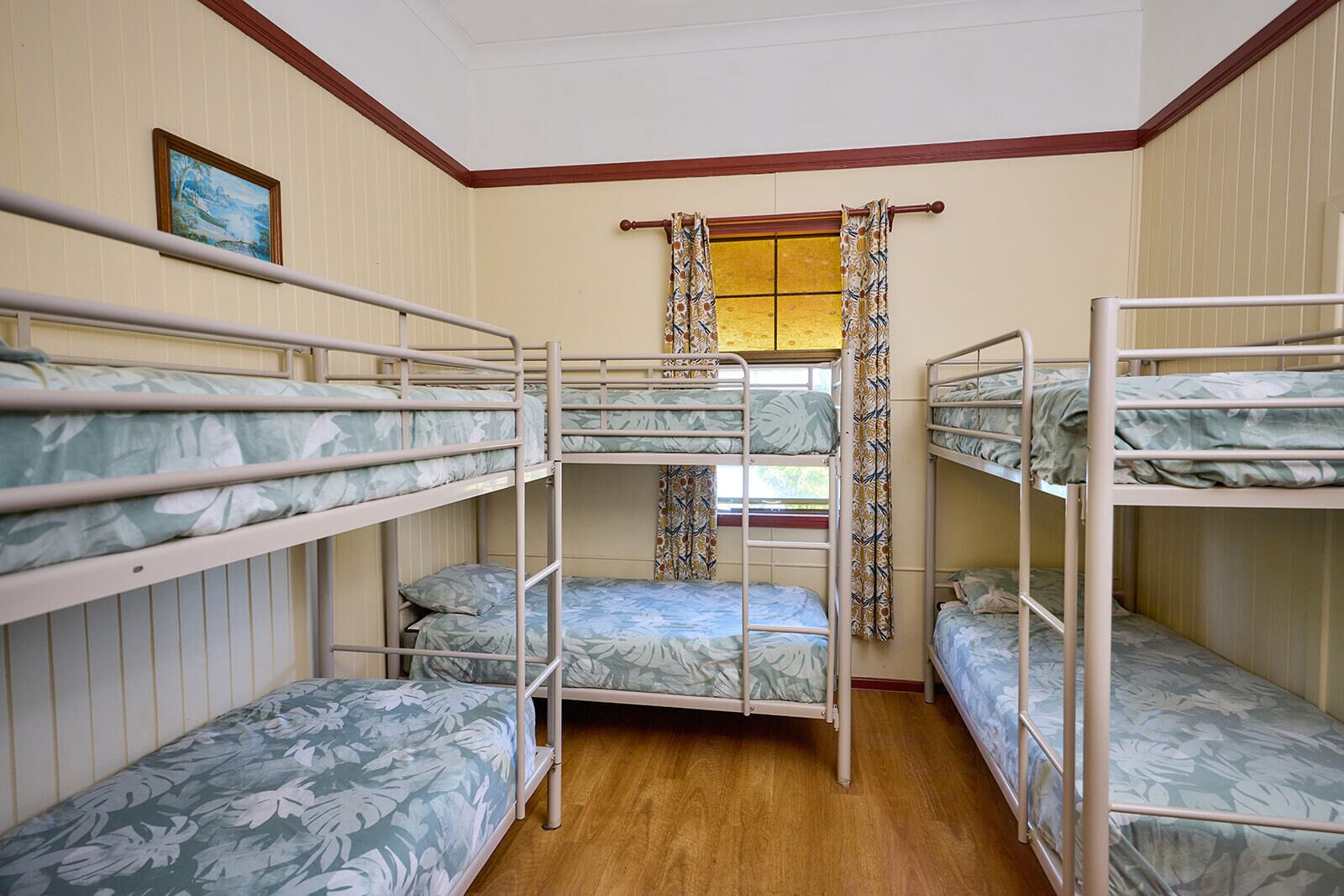 A dormitory room with bunk beds, wooden floors, a window with patterned curtains, and a framed picture on the wall.