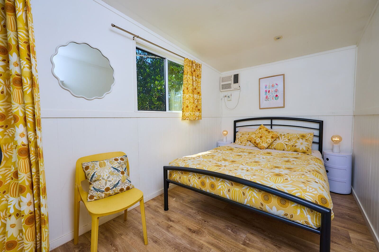 Un dormitorio con cama, una silla, un ventanal y un espejo instalado en la pared.