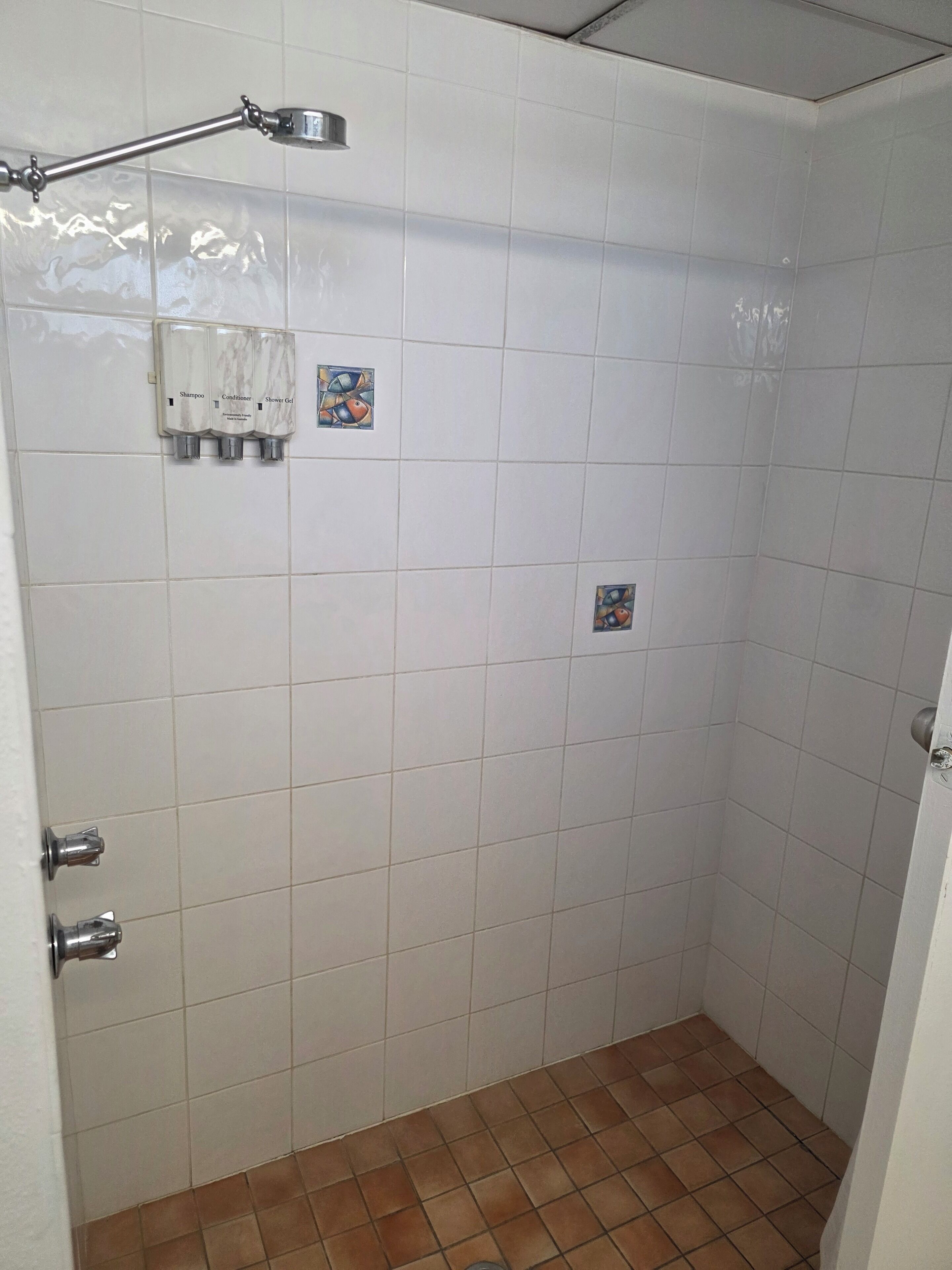 A shower area with white tiles, a showerhead, and three bottles of shampoo, conditioner, and shower gel.