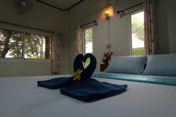 A hotel room with a bed, two windows with curtains, a bedside table with a vase of flowers, and a wall-mounted light fixture.