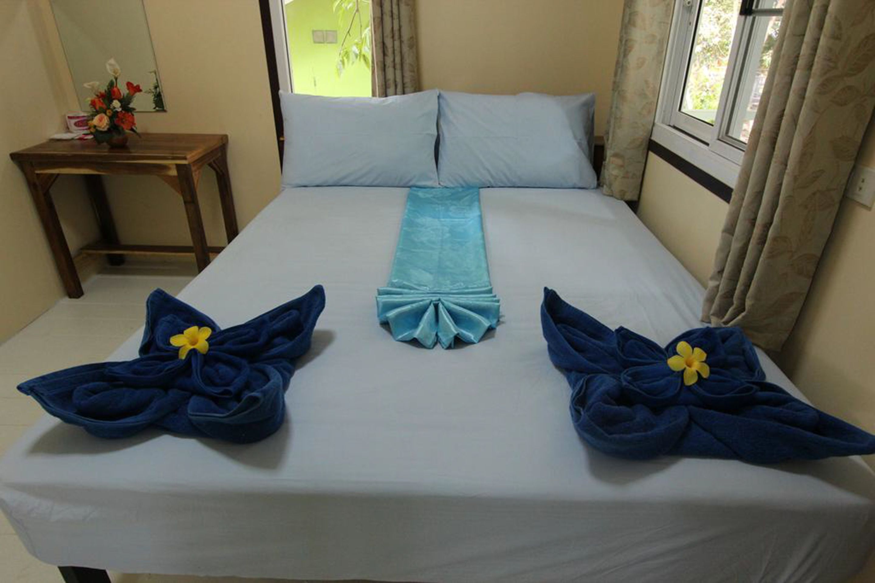 Una cama bien hecha con una colcha azul claro, dos toallas azules con flores amarillas y una mesita de noche de madera con un florero.