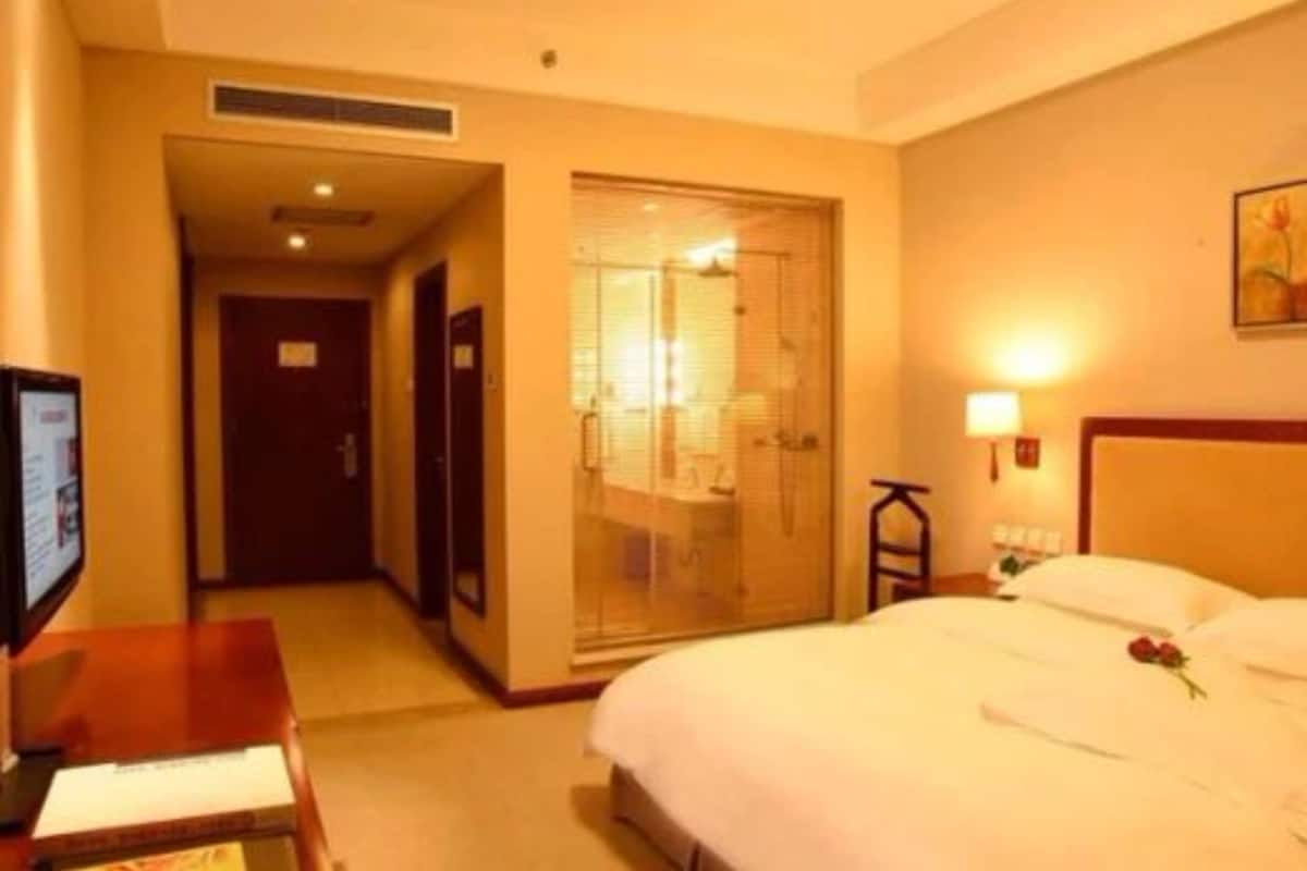Een hotelkamer met een bed, een bureau, een televisie, een badkamer met een glazen deur en een schilderij aan de muur.