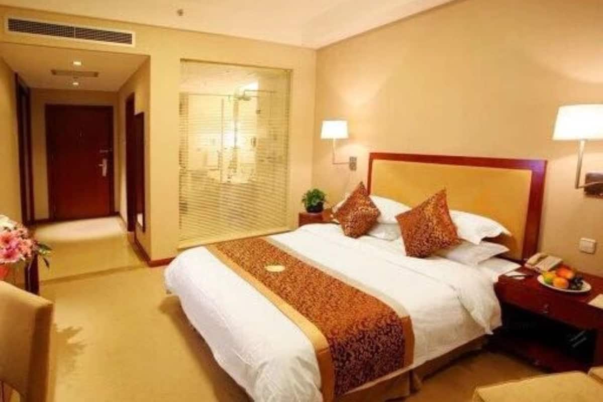 Een hotelkamer met een groot bed, twee bedlampen, een nachtkastje met een telefoon en een schaal fruit, en een deur naar een andere kamer.