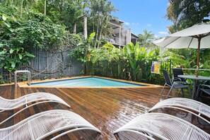A wooden deck with a pool, surrounded by lush greenery and white lounge chairs.