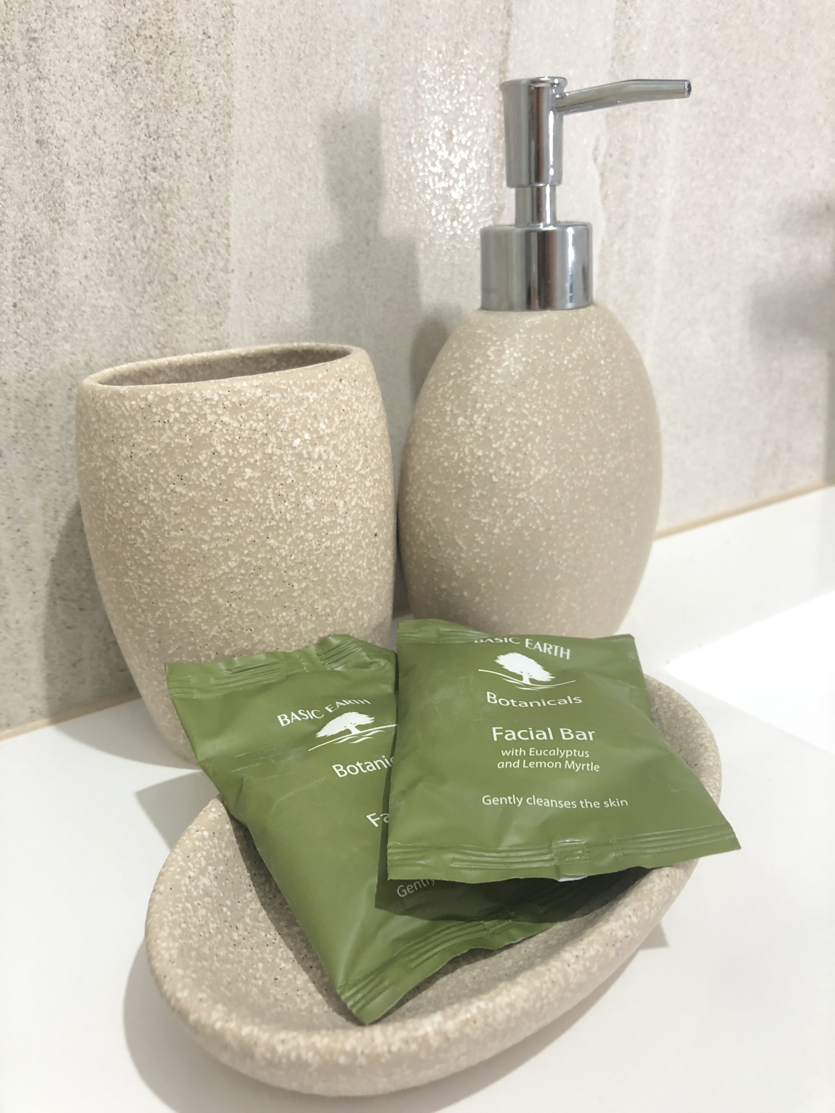 Un baño con un lavabo de color beige con textura, un grifo plateado y dos bolsas verdes con la etiqueta 'Basic Earth Botanicals Facial Bar'.