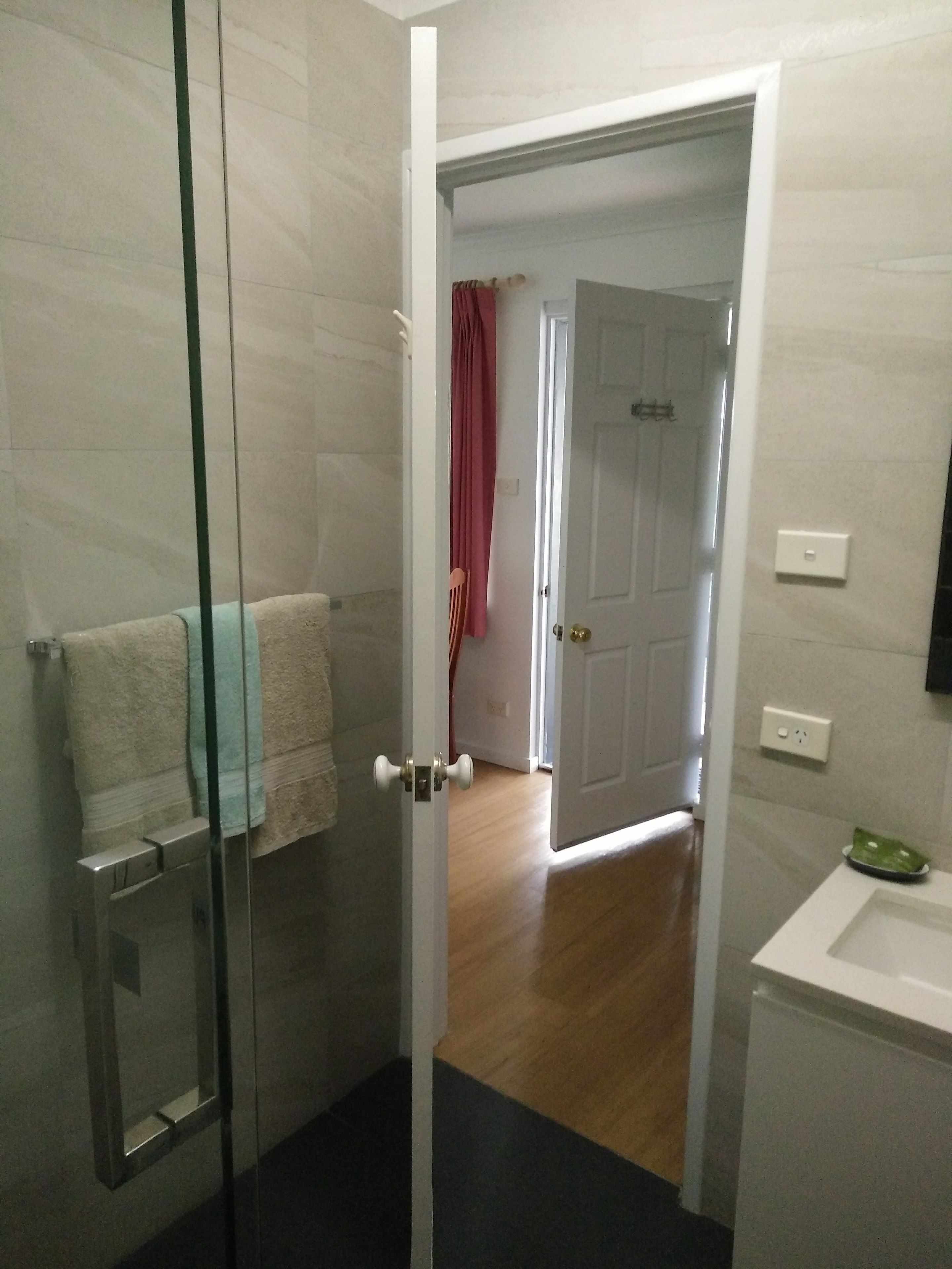 Un baño con mampara de vidrio, un perchero con toalla, un lavabo blanco y una puerta que da a otra habitación.