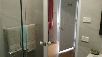 Un baño con mampara de vidrio, un perchero con toalla, un lavabo blanco y una puerta que da a otra habitación.