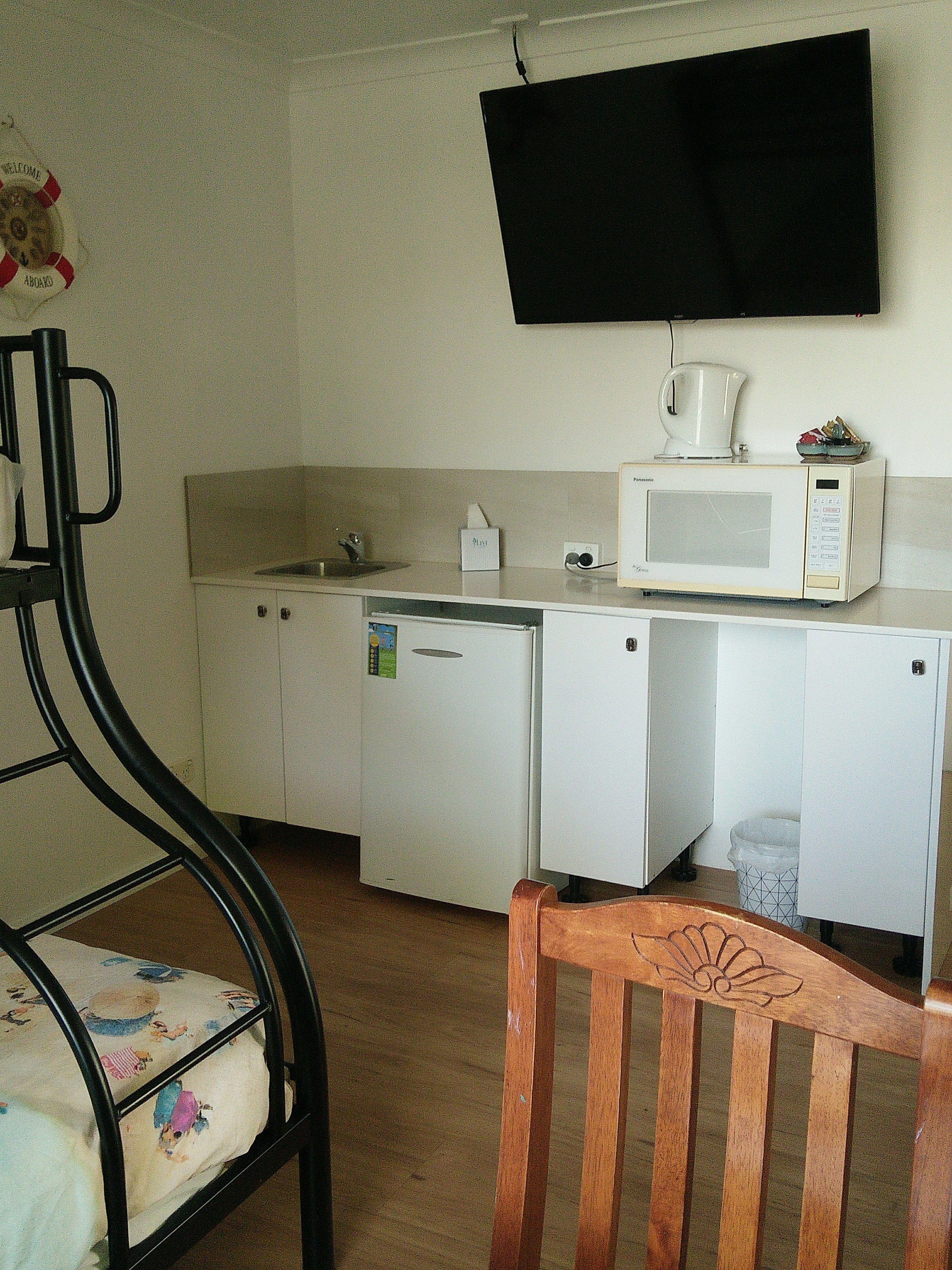 A compact kitchen with a microwave, coffee maker, and a small refrigerator. A wooden chair with a floral cushion is in the foreground.