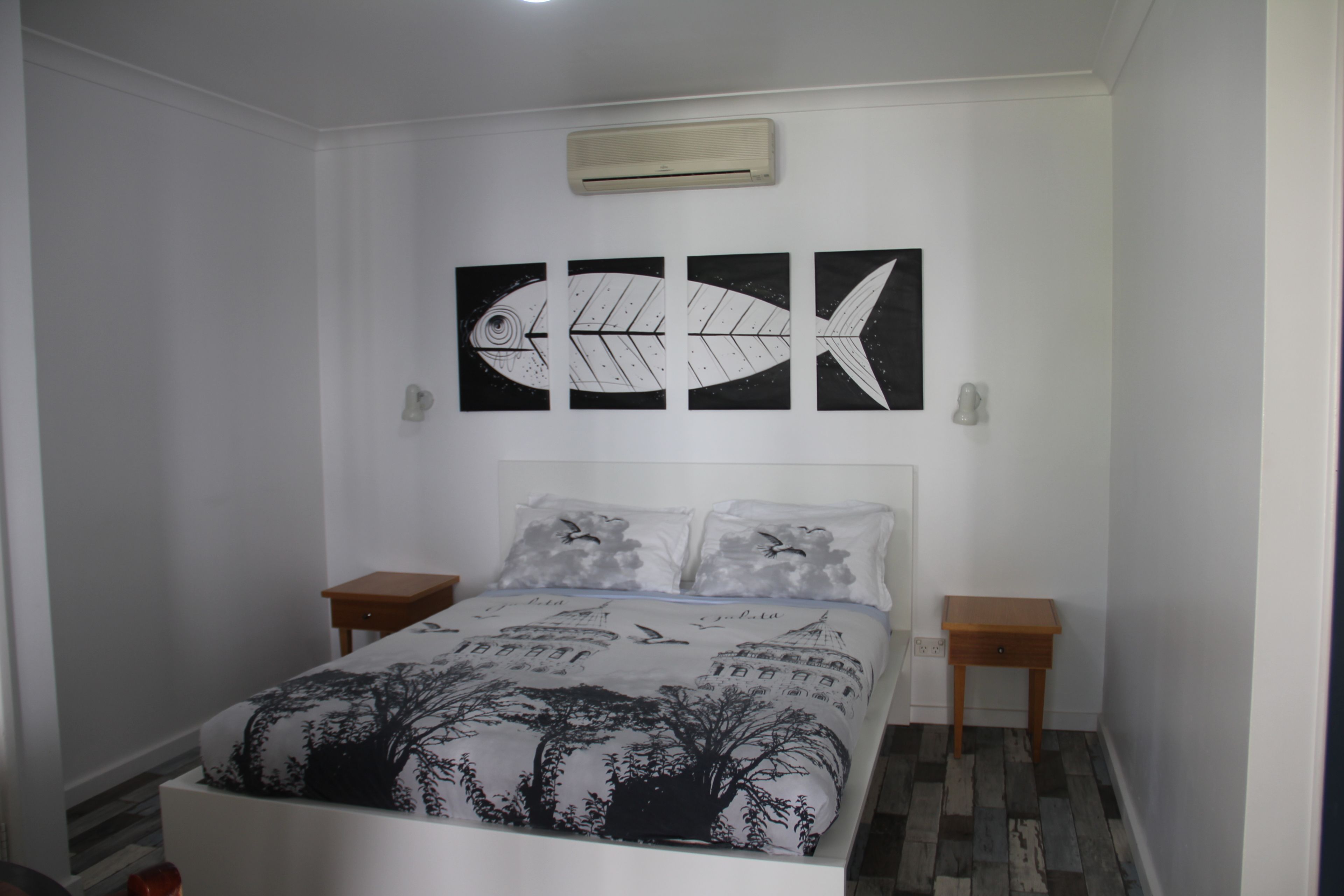Een slaapkamer met een bed, twee nachtkastjes, een wandairconditioner en drie kunstwerken met daarop vis.