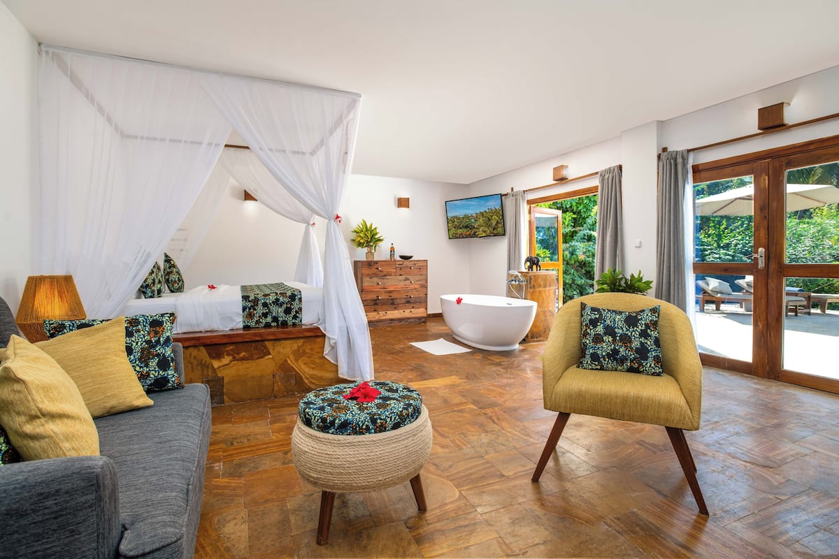 A bedroom with a canopy bed, a sitting area with a sofa and armchair, a wooden dresser, and a bathtub.
