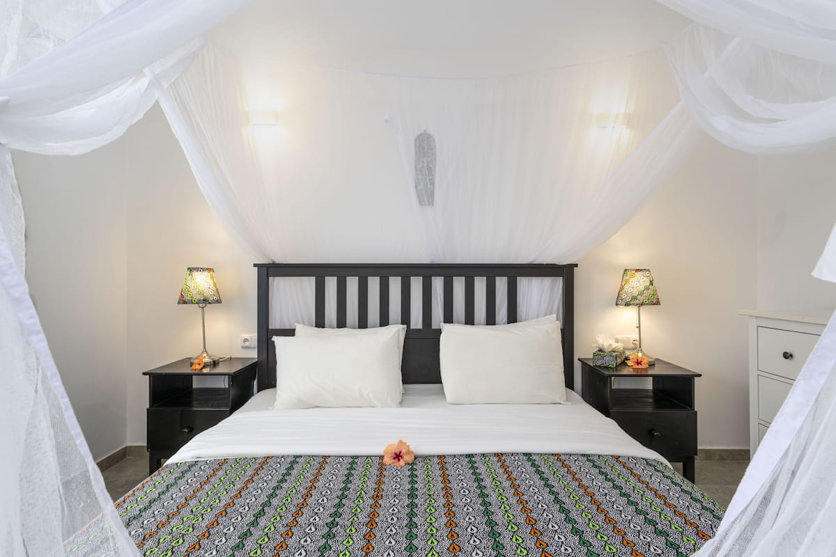 A bedroom with a canopy bed, two bedside lamps, a nightstand, and a bedside table with a lamp.