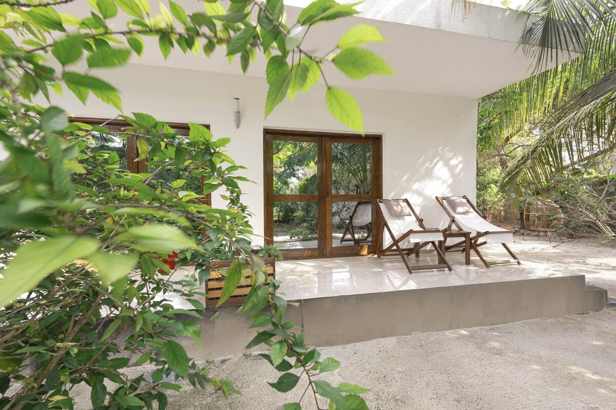A modern outdoor seating area with two lounge chairs and a concrete platform, surrounded by lush greenery and a large window.