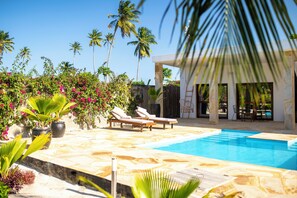 Un área de piscina con sillones reclinables, una piscina y plantas tropicales.