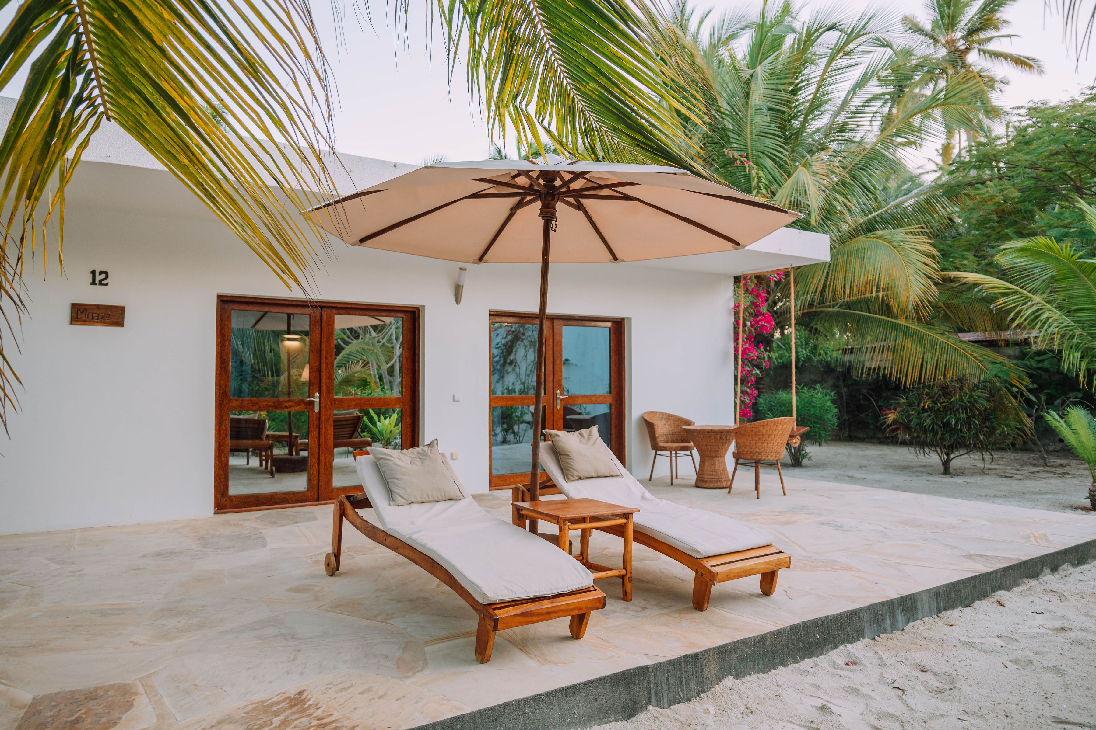 A sunlit outdoor lounge area with two wooden lounge chairs and a table, an umbrella, and a small garden with palm trees and flowers.