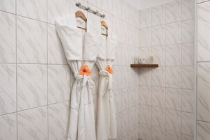 Un baño con baldosas de mármol blanco, dos albornoces blancos colgados en un perchero y una estantería de madera con artículos de aseo.