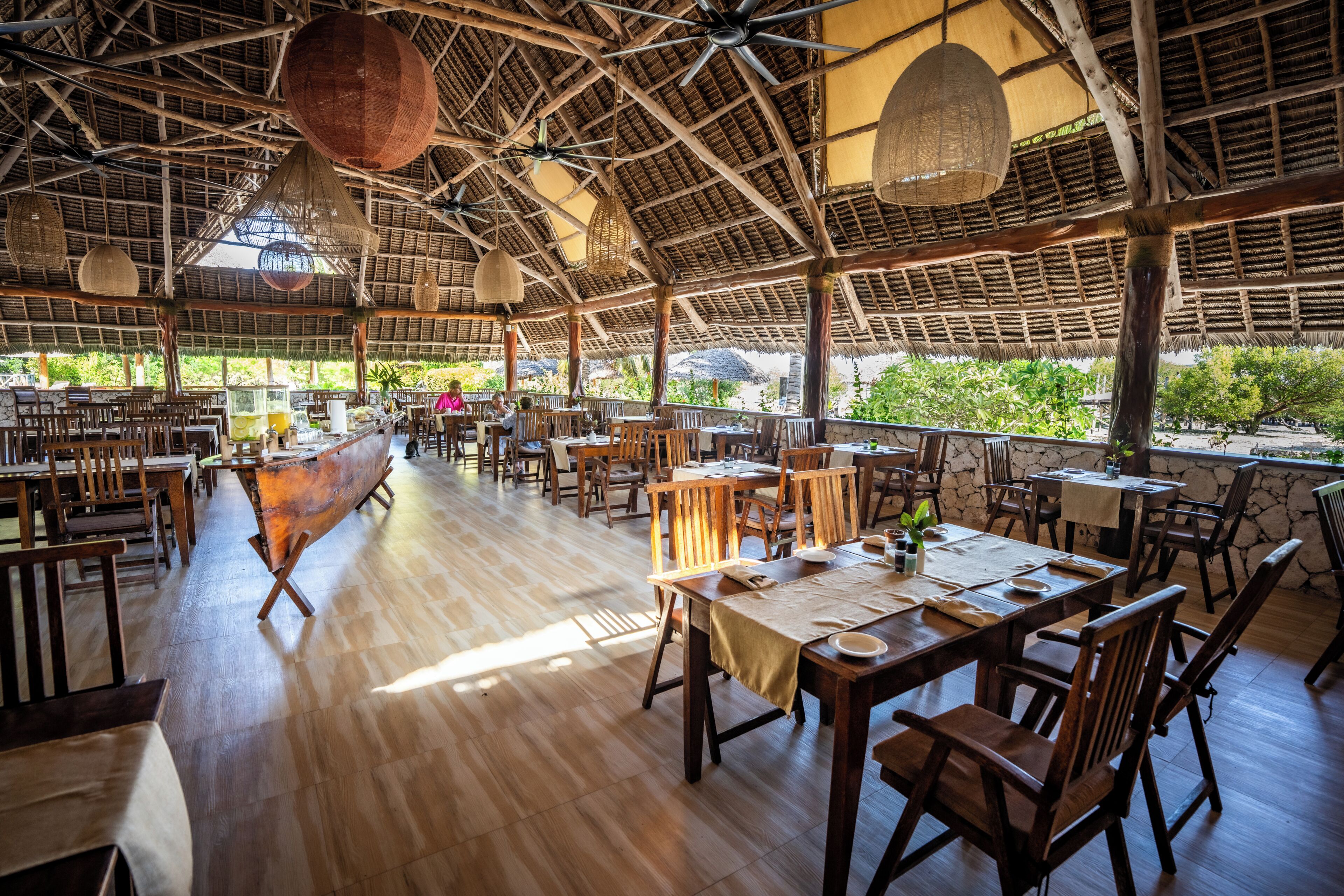 An open-plan restaurant with wooden furniture, a high ceiling with woven ceiling panels, and large windows overlooking greenery.