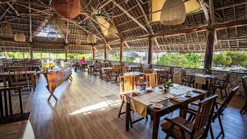 Un restaurante de planta abierta con mobiliario de madera, un techo alto con paneles tejidos y grandes ventanales con vistas a la vegetación.