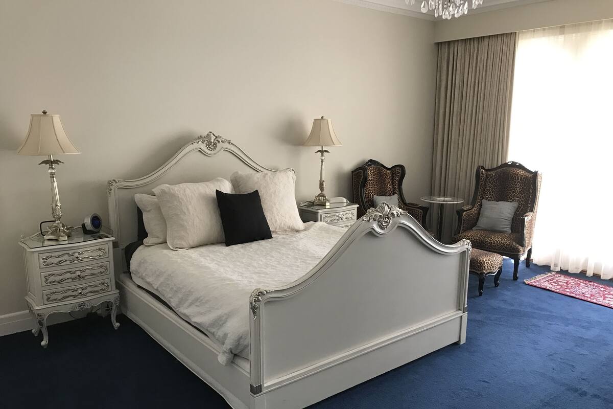 A classic four-poster bed with ornate detailing, two bedside tables with lamps, a leopard print armchair, and a blue carpet.