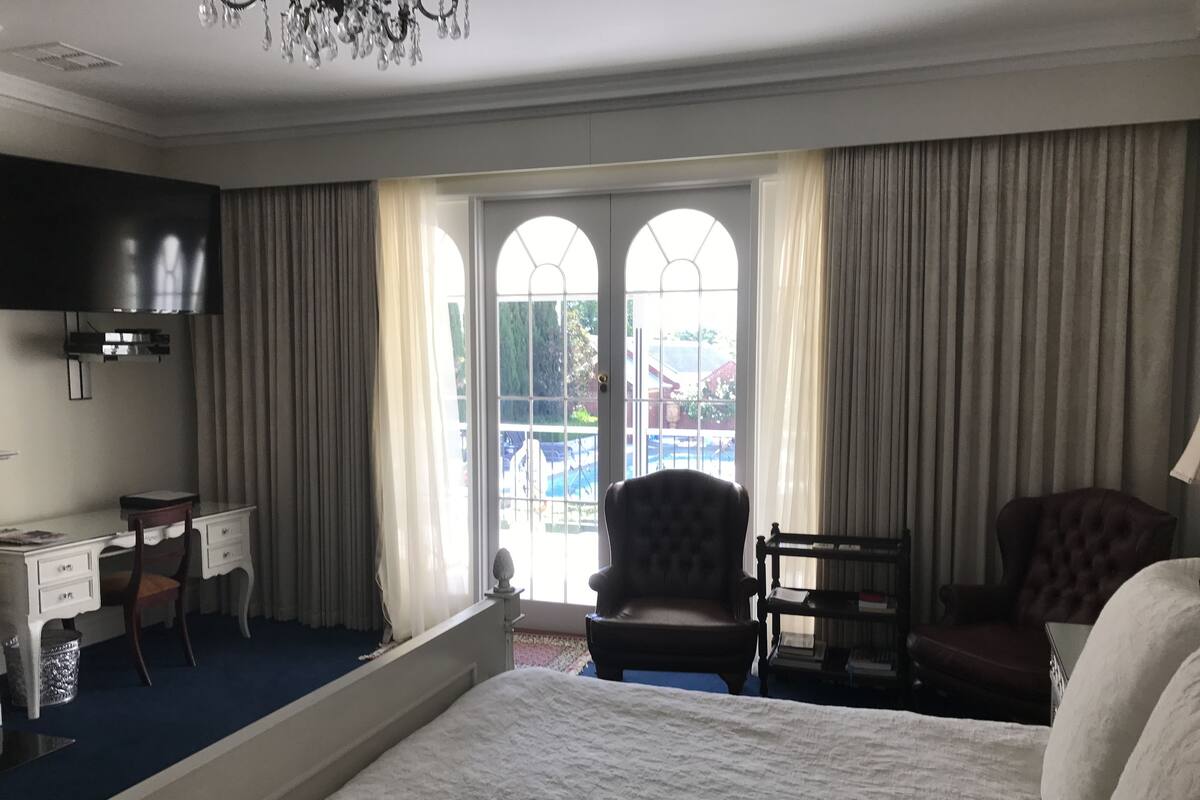 A hotel room with a bed, a desk, two armchairs, a chandelier, and a large window with curtains.