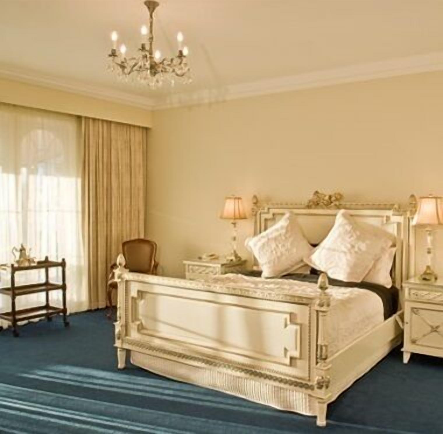 A classic four-poster bed with ornate detailing, a chandelier, and a nightstand with a lamp.