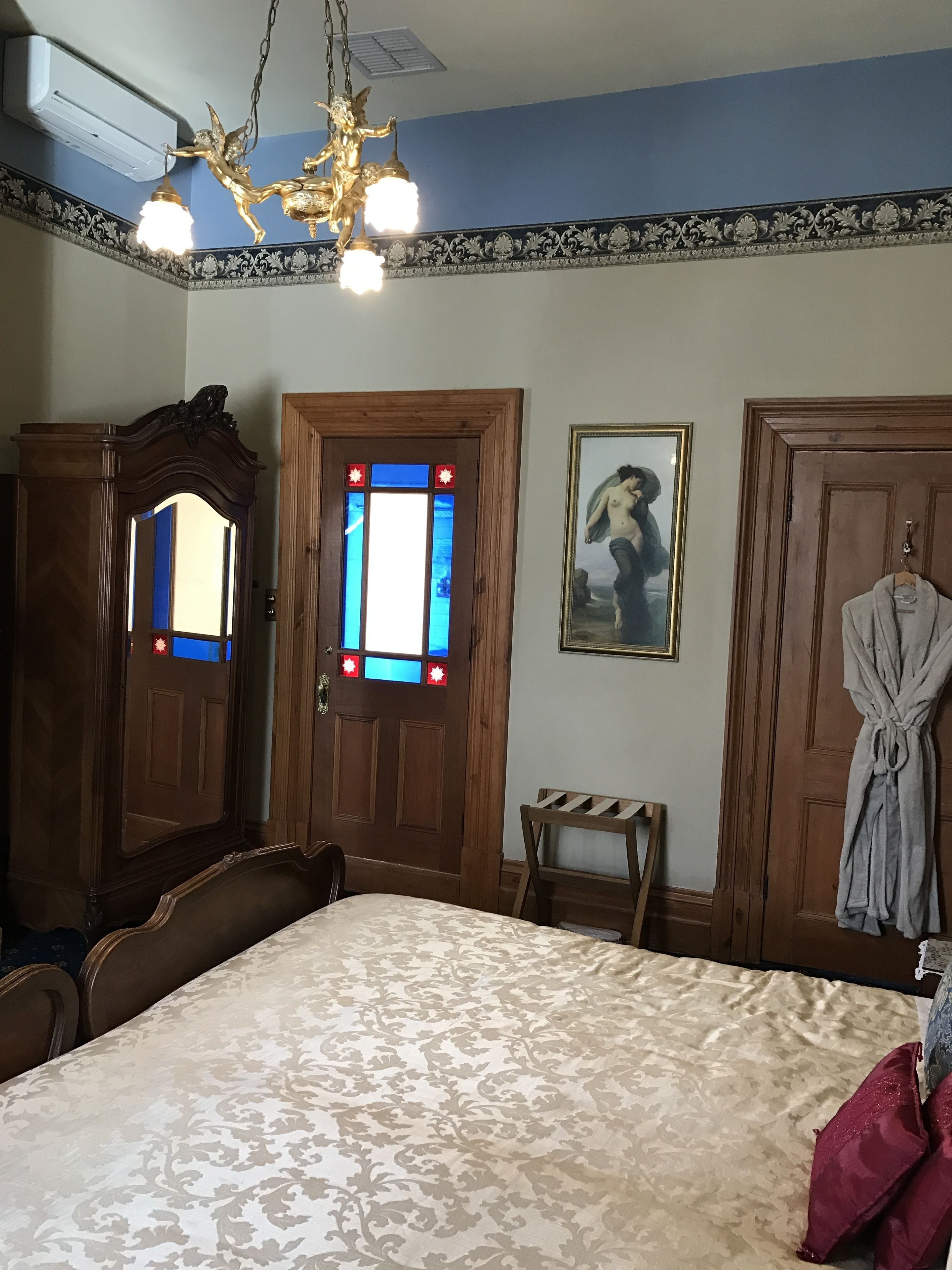 A bedroom with a large bed, a wooden wardrobe, a small table, a robe hanging, and a chandelier.