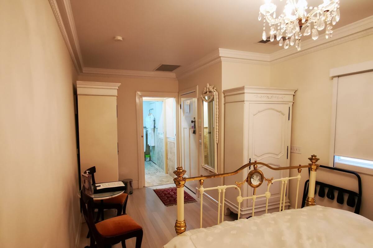 A bedroom with a bed, a desk, and a chair. There is a mirror and a chandelier.