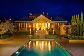 Un edificio grande y bien iluminado con una piscina frente a él, rodeado de jardines ajardinados y árboles altos.