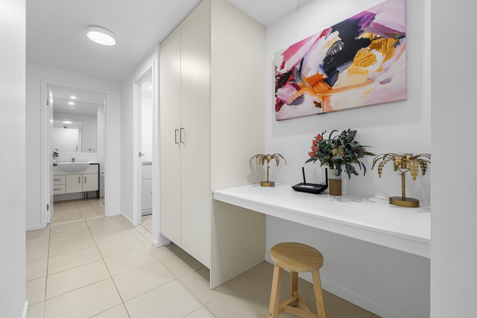 Een moderne keuken met witte kastjes, een wit aanrechtblad en een schilderij erboven. Twee potplanten staan op het aanrecht.