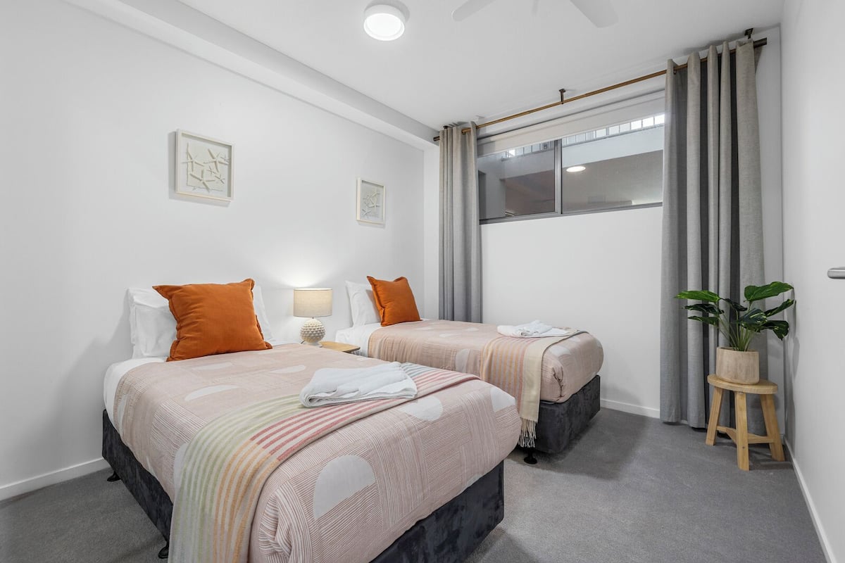 Habitación de hotel con dos camas, una ventana con cortinas, un taburete de madera con una planta y un ventilador de techo.