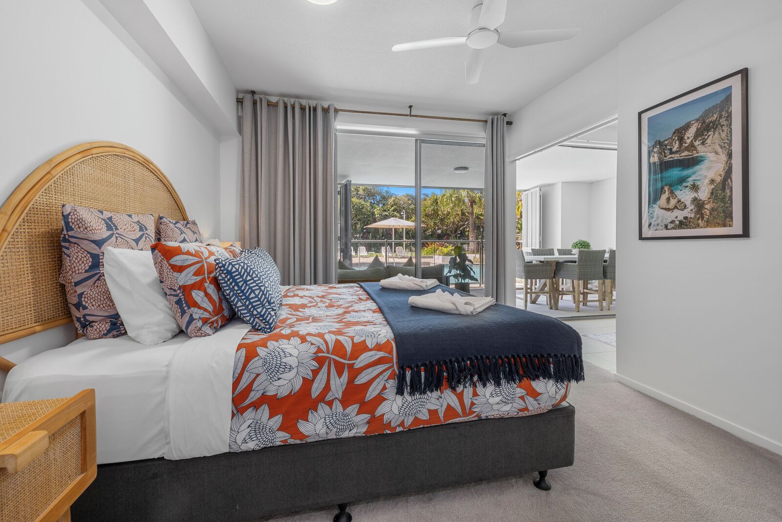 A bedroom with a bed, a ceiling fan, a large window with curtains, and a view of the outdoors.