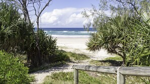 Ein Sandweg am Strand führt zum Meer, umgeben von üppiger Vegetation und einem Holzzaun.