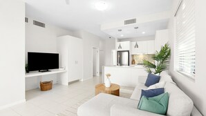 Ein modernes Wohnzimmer mit Flachbildfernseher, einer Couch mit Kissen, einem Couchtisch und einer Küche im Hintergrund.