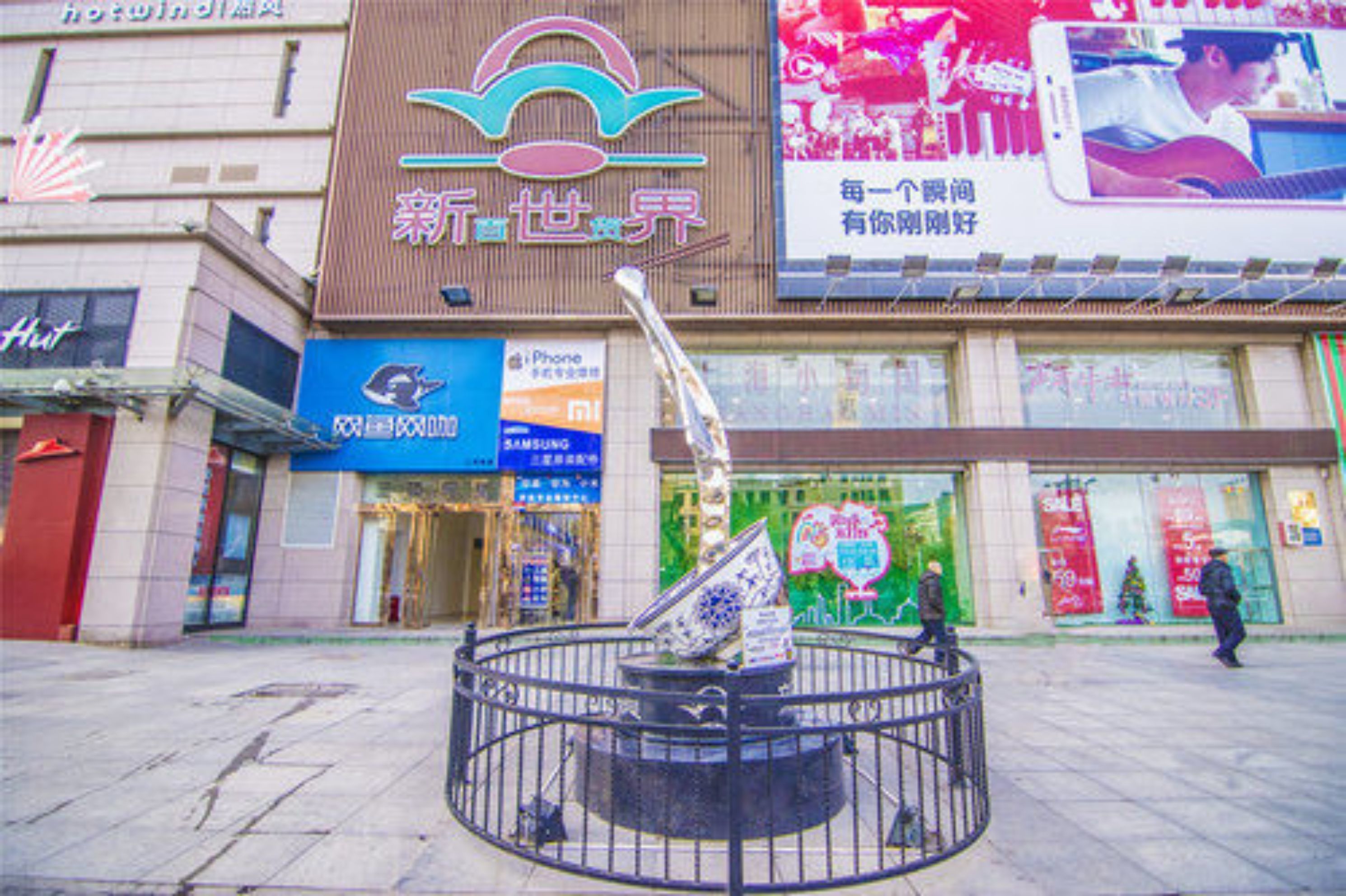 A shopping mall with a central fountain, various storefronts, and a large digital advertisement.