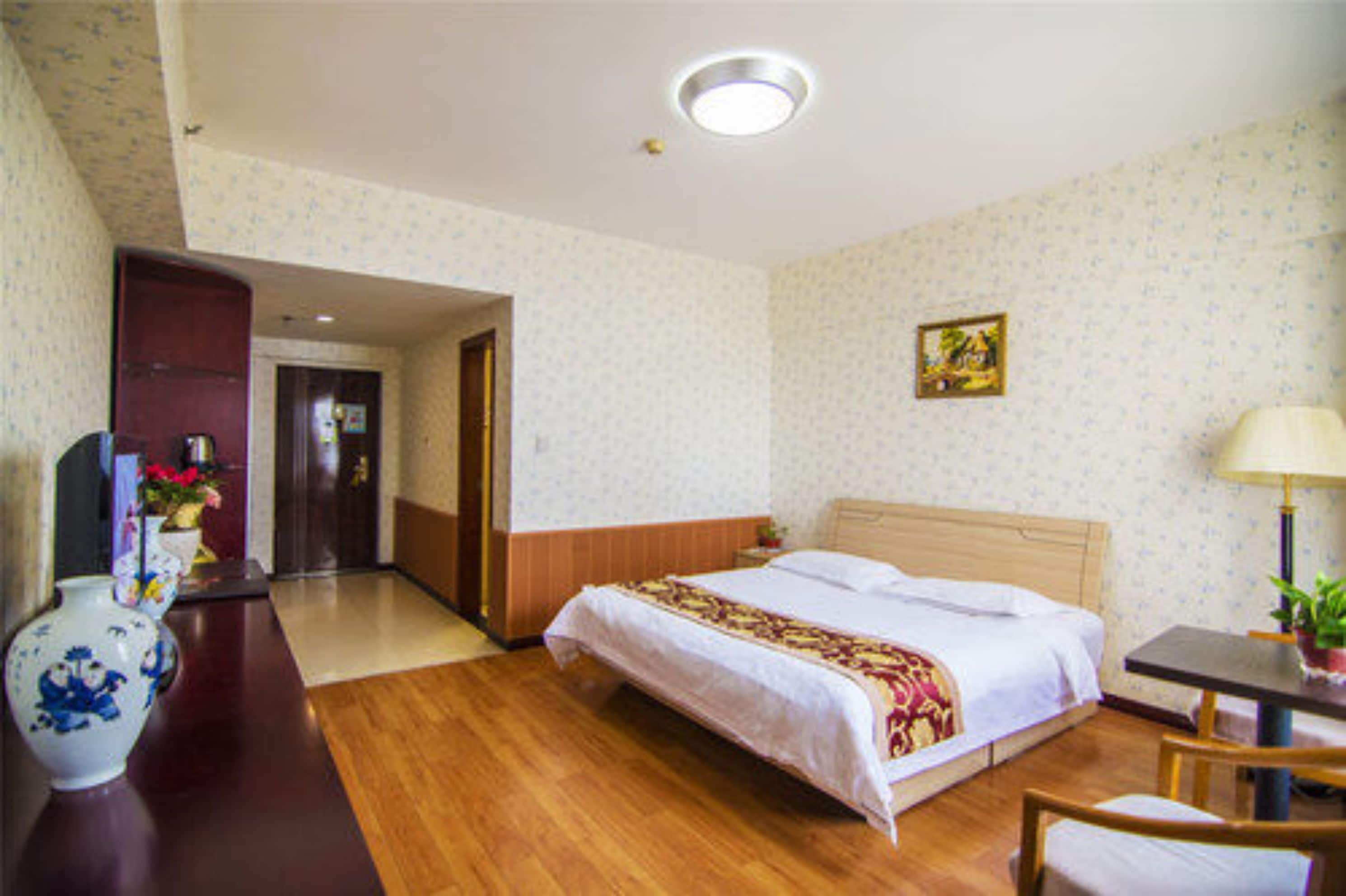 Una habitación de hotel con piso de madera, una cama con cubrecama blanco, un escritorio con lámpara y un florero con flores.
