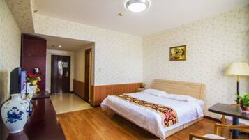 Una habitación de hotel con piso de madera, una cama con cubrecama blanco, un escritorio con lámpara y un florero con flores.