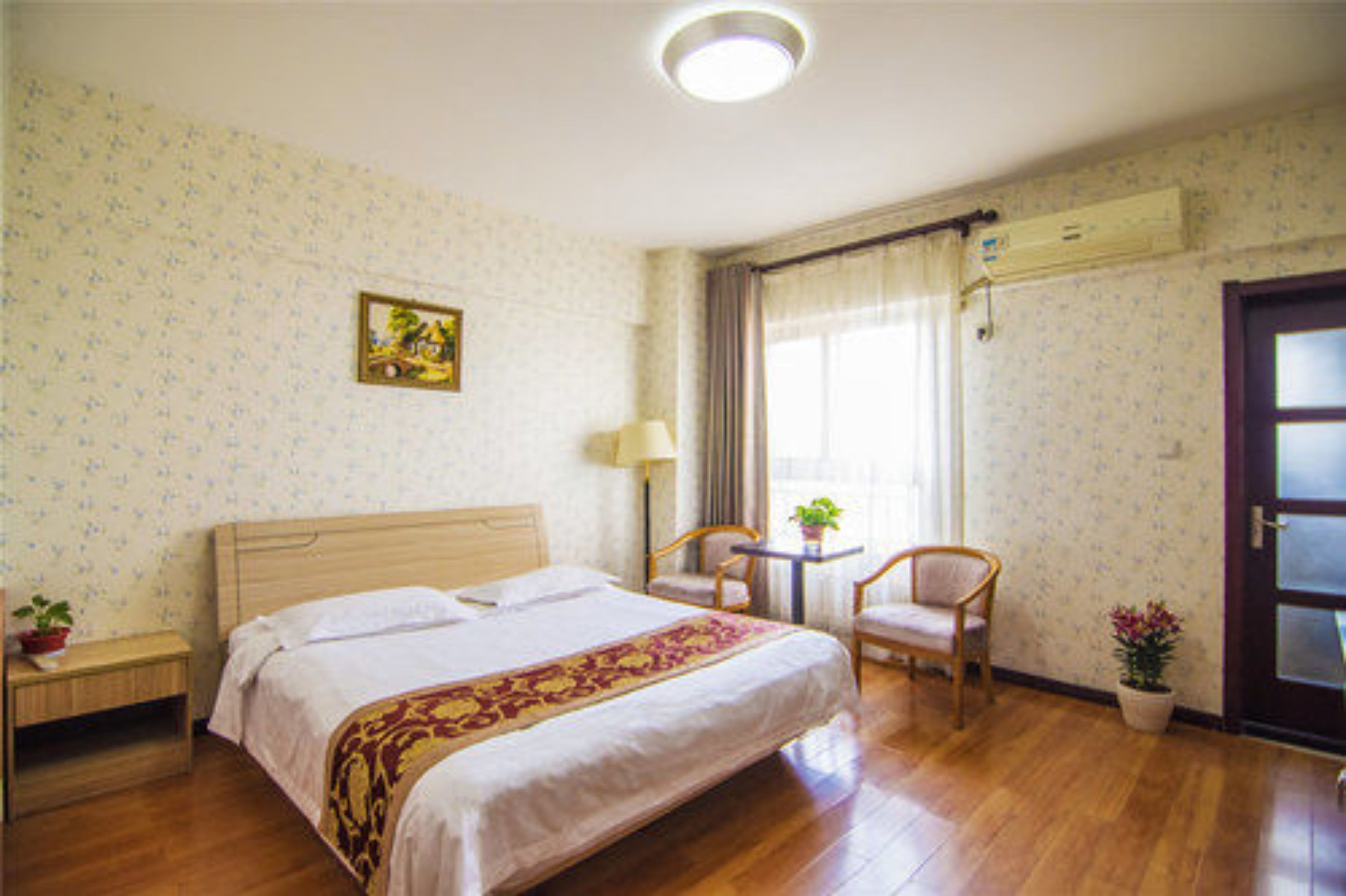 A hotel room with a large bed, a desk with a chair, a small table with a plant, and a door with a frosted glass panel.