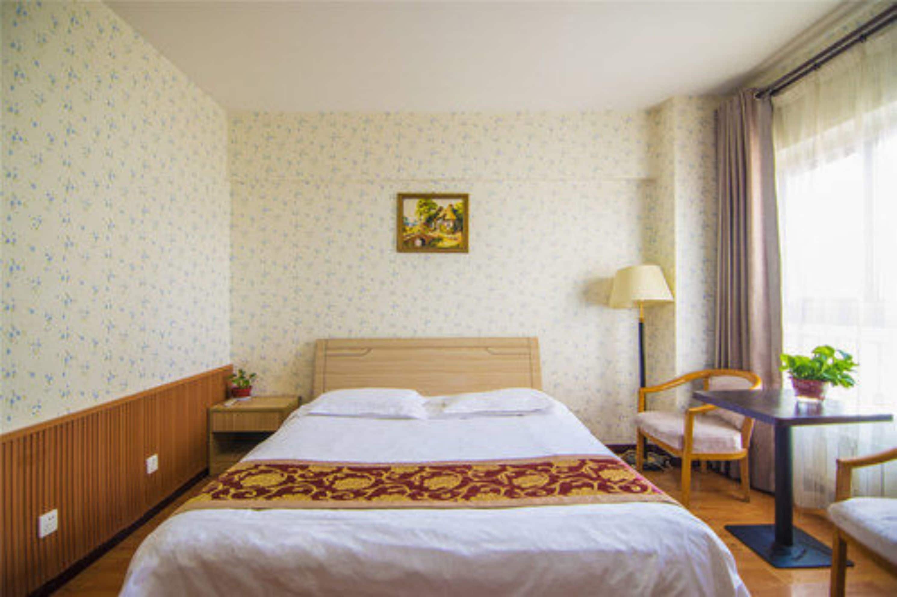 A hotel room with a large bed, a bedside table with a lamp, a chair, and a small table with a plant.