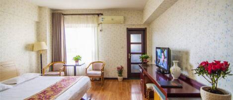 A hotel room with a bed, two chairs, a TV, a small table, and a potted plant.