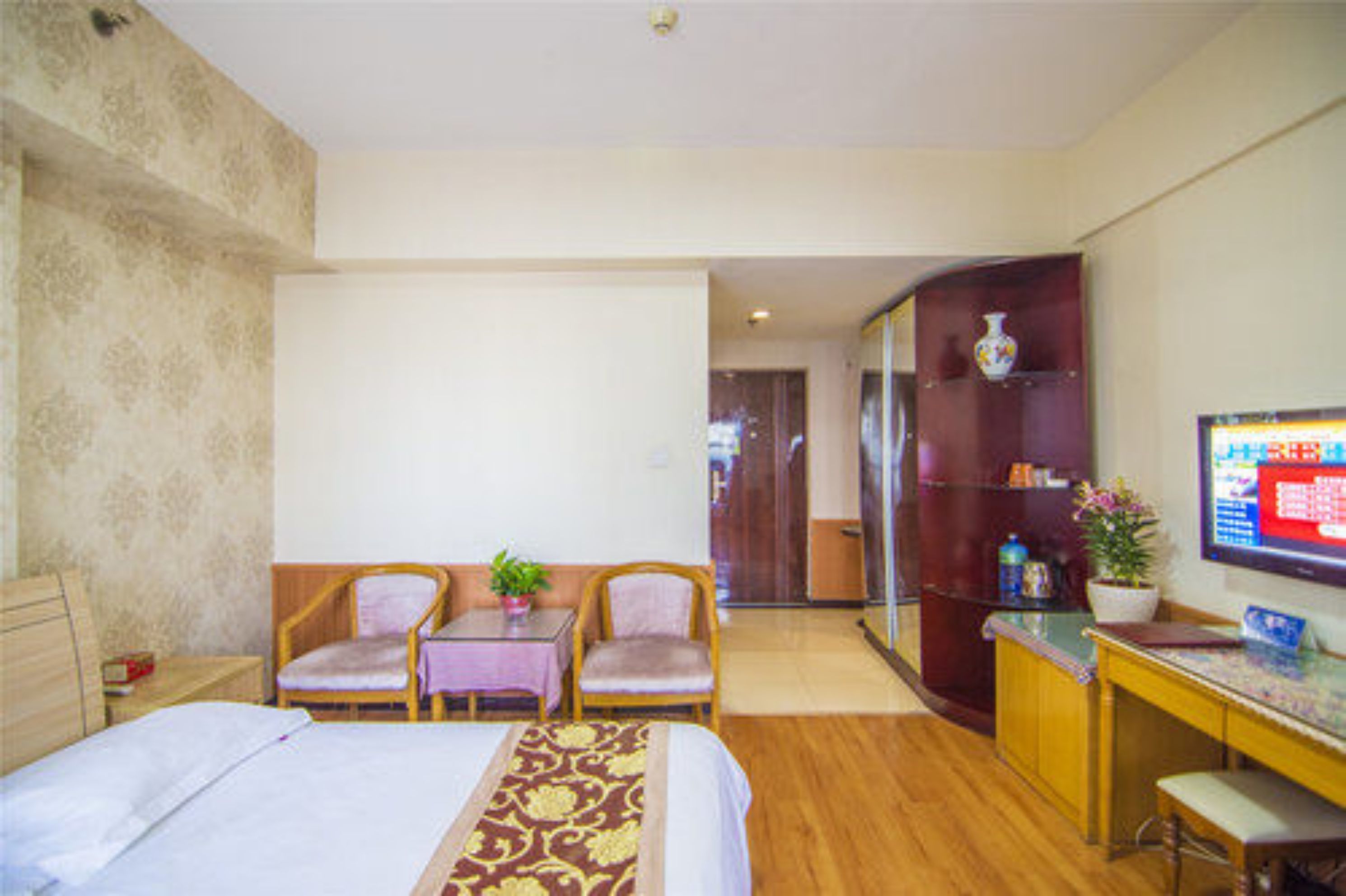 A hotel room with a bed, two chairs, a small table, a desk, a TV, and a cabinet with decorative items.