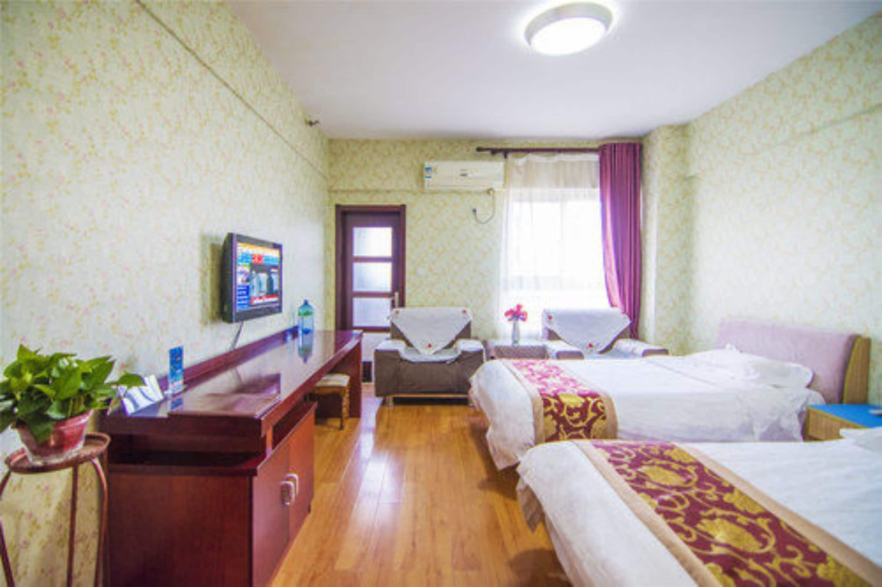 A hotel room with two beds, a desk with a computer, a chair, a small table with a vase, and a potted plant.