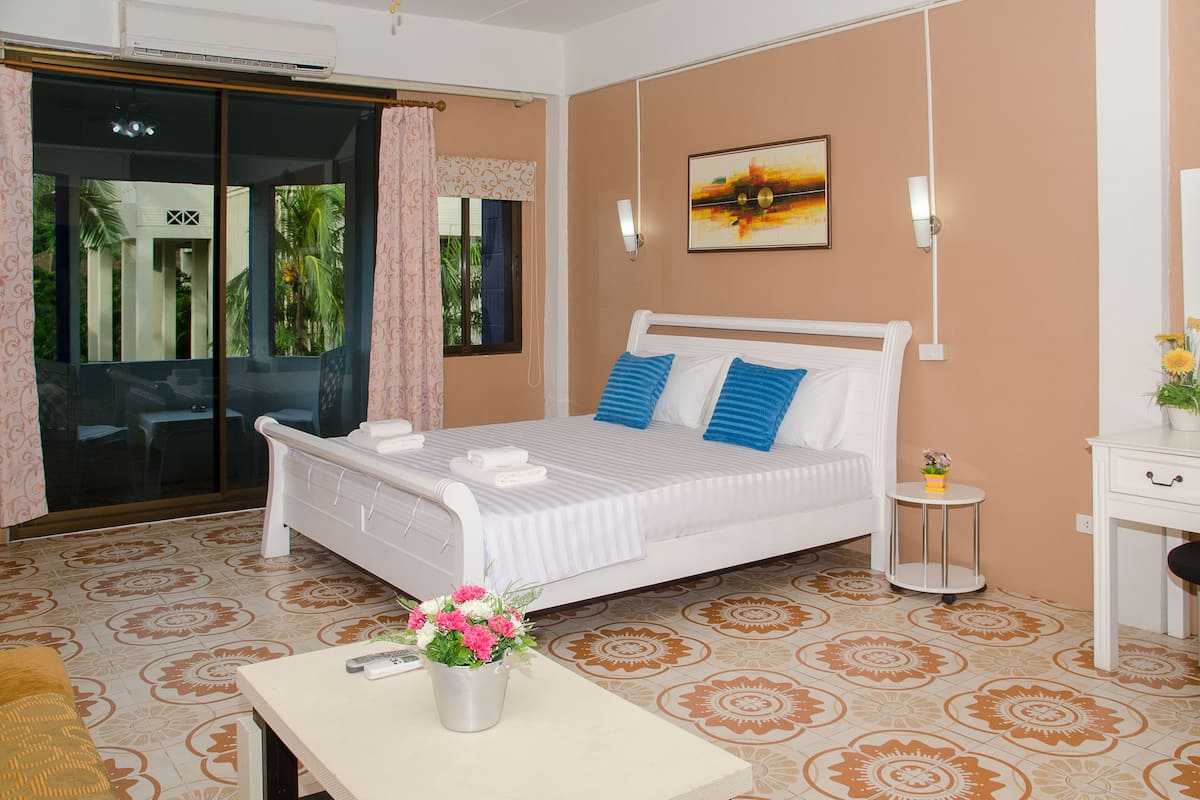 A bedroom with a bed, a small table with a vase of flowers, a bedside table, a lamp, and a view of the outside through a sliding door.