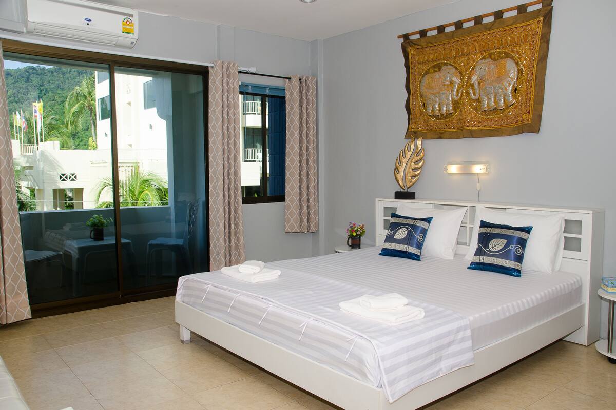 A hotel room with a large bed, a sliding glass door to the balcony, and a decorative elephant tapestry on the wall.
