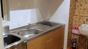 A kitchen with two stainless steel sinks, wooden cabinets, a white countertop, a window, and a fire extinguisher mounted on the wall. - OCHO Guest House - Hostel (Sapporo)