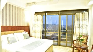 A hotel room with a large bed, a balcony with a city view, a small table with chairs, and a vase with flowers. - Interservice Hotel (Kampala)