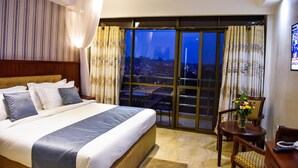 A hotel room with a large bed, a balcony with a city view, and a small table with a vase of flowers. - Interservice Hotel (Kampala)