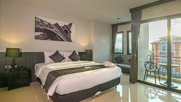 A modern hotel room with a large bed, two bedside tables, a nightstand, a balcony with a chair, and a cityscape view. - The Rich Sotel (Krabi)