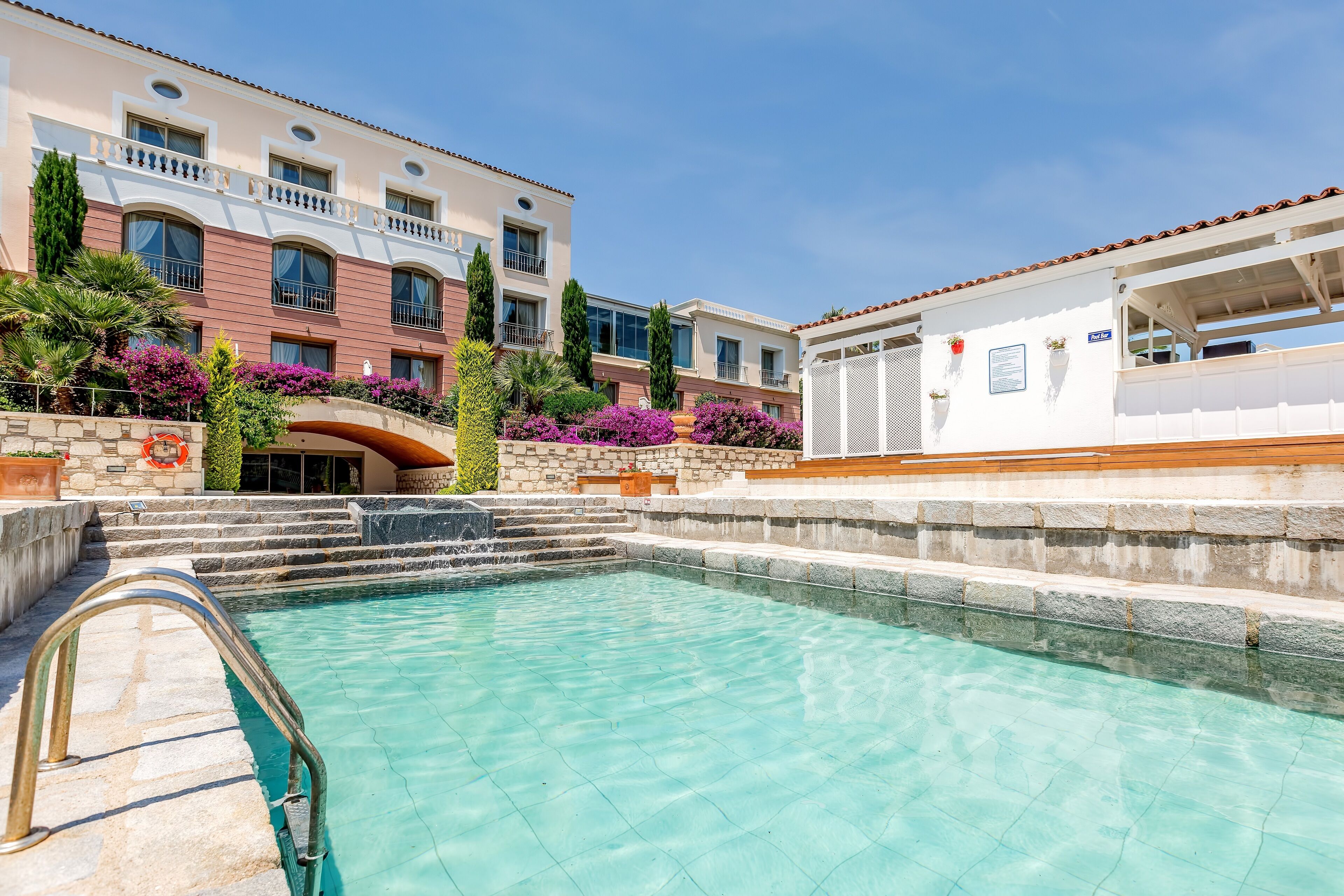A pool area with a clear blue pool, stone steps leading into the pool, and a building with balconies and windows in the background.