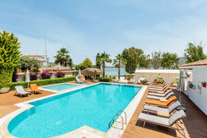 Una piscina rectangular con cubierta de madera, rodeada de sillas de descanso y sombrillas, con vista al mar y follaje en el fondo.