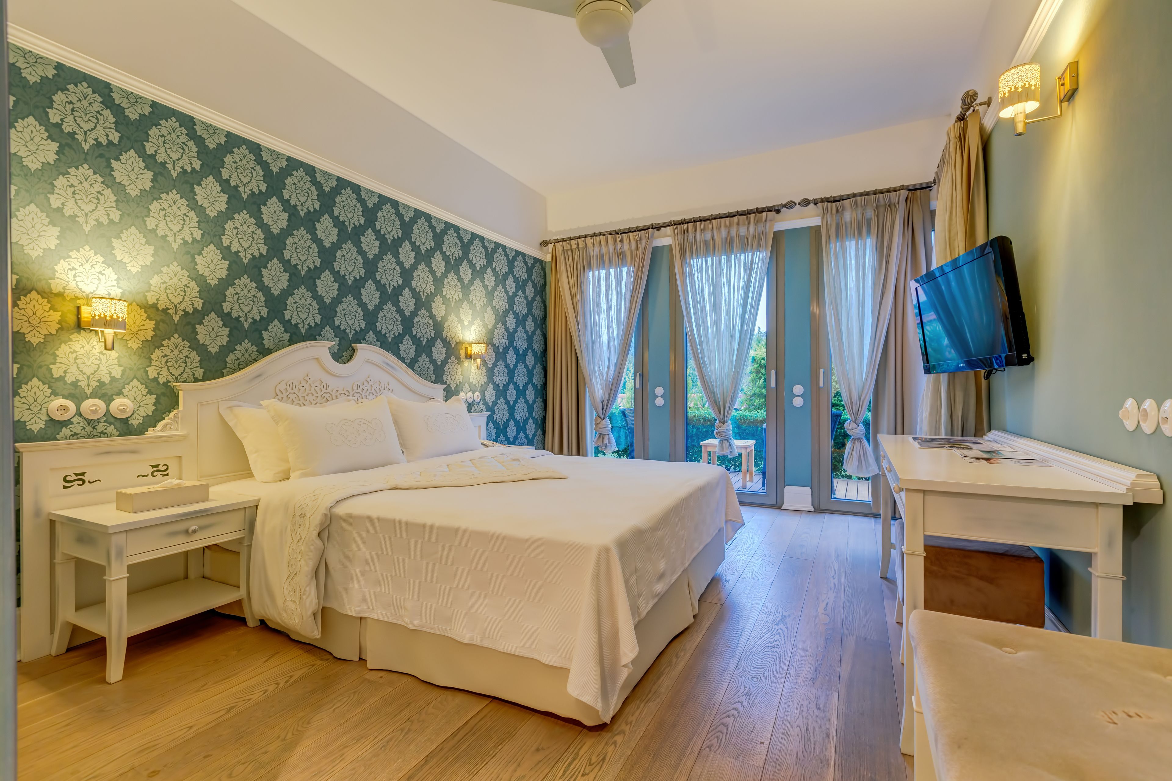 A bedroom with a bed, bedside tables, a desk, a television, and a view of the outdoors.